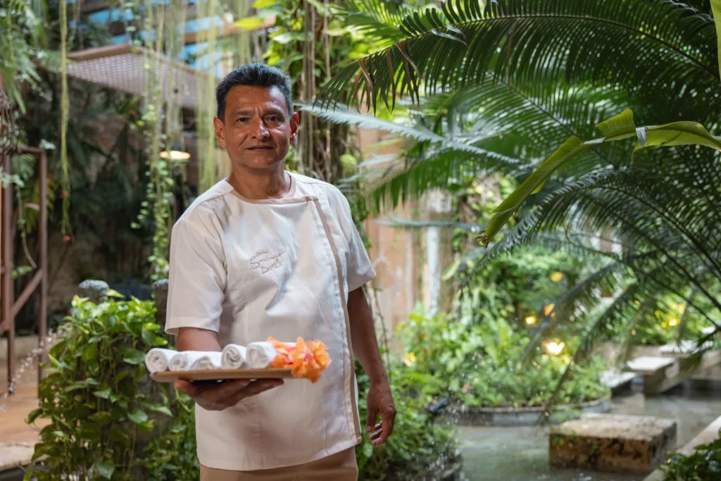 Integrante del equipo de la casa recibe a los huéspedes con una bandeja de toallas y flores en el jardín